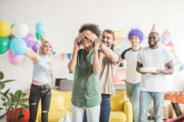 Happy friends covering eyes of young woman and greeting her with birthday cake  on surprise party