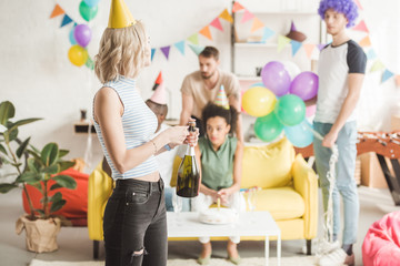 Blonde girl opening champagne in front of partying friends