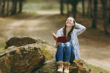 Young relaxing smiling beautiful woman in casual clothes sitting on stone studying reading book in big city park or forest on green background. Student learning, education. Lifestyle, leisure concept.