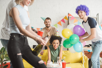 Cheerful young people looking at woman opening champagne at birthday party