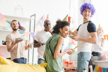 Young people in party hats celebrating birthday with drinks in cozy room