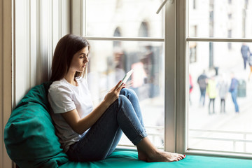Beautiful smiling brunette girl wears white t-shirt using tablet computer sitting on the windwsill