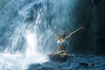 attractive woman sitting at rock in yoga pose for spiritual relaxation serenity and meditation at stunning beautiful waterfall and rain forest in Bali
