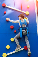Fototapeta premium Every mountain top is within reach if you just keep climbing! Little boy having great time, while climbing a rock wall indoors.