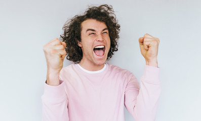 Portrait of positive successful handsome young male wears pink sweater, clenches fists in joy, opens mouth widely as exclaims with happiness, isolated over white studio background. Advertisement
