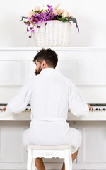 Man sleepy in bathrobe sit in front of piano musical instrument in white interior on background, rear view. Man in bathrobe enjoys morning while playing piano. Talented musician concept.