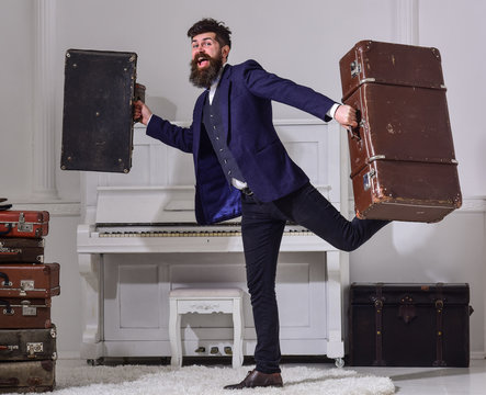 Man, traveller with beard and mustache with luggage, luxury white interior background. Hotel service concept. Macho elegant on cheerful face walks near pile of vintage suitcase, holds suitcases.