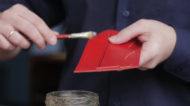 Leathersmith is applying glue on a leather parts of the zipper with a special brush. He works at the table in the workshop.