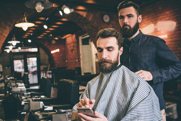 Bearded guy is sitting in chair and getting ready for procedure. He is looking at the phone. He is very concentrated. Hairdresser is ready to start procedure.