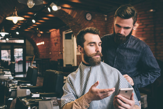 Bearded guy is showing to hairdresser a picture on the phone. Hairdresser is looking at the phone with serious sight.
