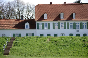 Wohnhaus hinter dem Deich / Das Dach sowie die Fensterreihe eines Wohnhauses hinter einem grünen Deich.