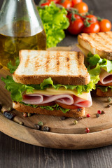Close-up photo of a club sandwich. Sandwich with meet, prosciutto, salami, salad, vegetables, lettuce, tomato, onion and mustard on a fresh sliced rye bread on wooden background. Olives background.
