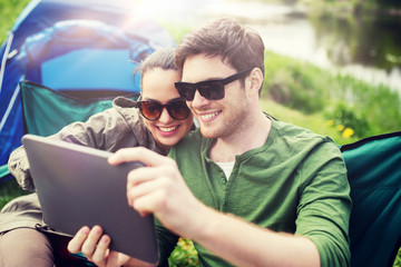 travel, hiking, technology, tourism and people concept - smiling couple with tablet pc computer sitting on chairs at camping tent