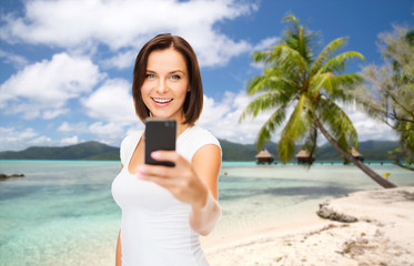 travel, tourism, summer holidays and vacation concept - happy young woman taking selfie by smartphone over exotic tropical beach with palm tree and bungalow sheds background in french polynesia