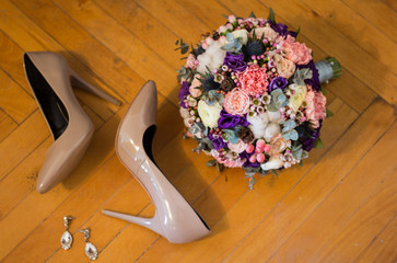 Beige high heels pumps shoes are lying on wooden old floor. Bridal bouquet and fashion jewelry earrings. Colorful flowers with eucalyptus, roses and purple eustoma. Wedding details.