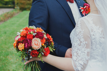Plus size wedding couple is standing and hugging outside. Curvy bride is holding beautiful colorful bouquet with orange, red and pink peonies and roses. Bride and groom in summer green love story.