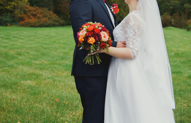 Plus size wedding couple is standing and hugging outside. Curvy bride is holding beautiful colorful bouquet with orange, red and pink peonies and roses. Bride and groom in summer green love story.