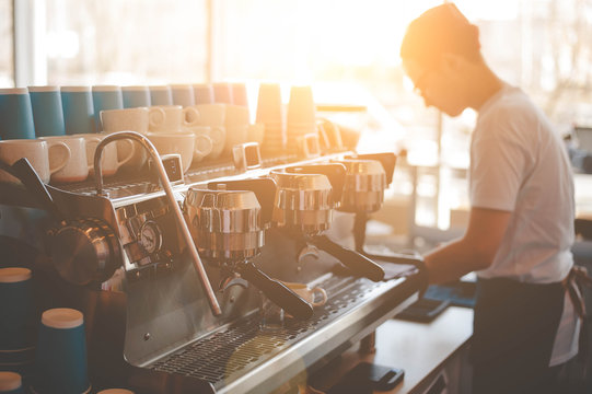 A young Barista guy stands behind a coffee machine and prepares fresh espresso, and the window shines a bright sun. Side view with copy space for your text.
