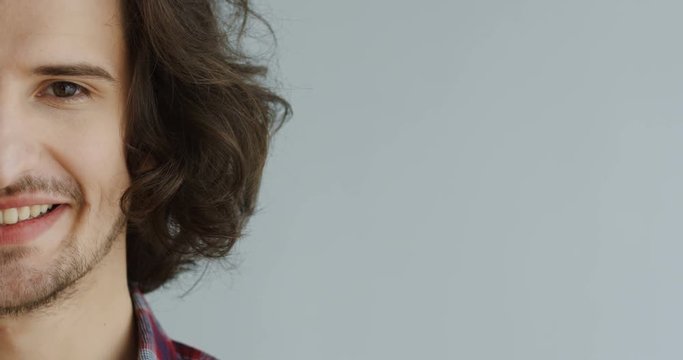 Close up of the half male face with long hair looking in the camera and then smiling on the white wall background. Inside