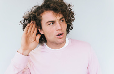 Closeup portrait of amazed eavesdropping male with curly hair placing hand on ear asking someone to speak up, isolated over white background. Handsome man which overhears conversation. Copy space