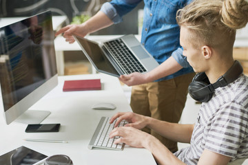 Pensive talented young web designer with hair bun typing on keyboard and looking at laptop screen while working with colleague in office