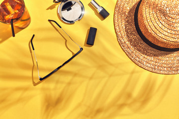 Straw beach woman's hat, beverage with ice in a glass and sun glasses on yellow background with shadow from a palm leaf.
