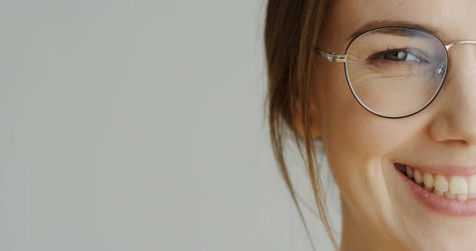 Close up of the half female facen in glasses with long fair hair looking in the camera and then smiling on the white wall background. Indoor