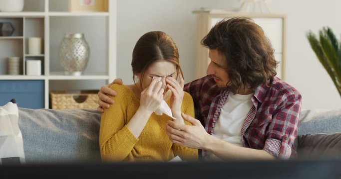 Young attractive couple sitting on the sofa, watching a melodrama movie and man calming down a woman as she crying. Indoor
