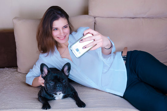 young girl taking selfie with dog. black active French bulldog lay on the couch. happy, smiling dog lover, dogman  hug,hold pet. cute caucasian teenager in modern, white shirt. woman looking to phone