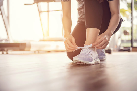 Woman Tying Running Shoes On Black Floor Background In Gym With Sunlight. Copy Space.