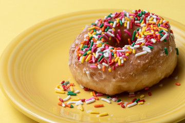 Close up of glazed doughnut with colorful sprinkles on a yellow plate with yellow background.