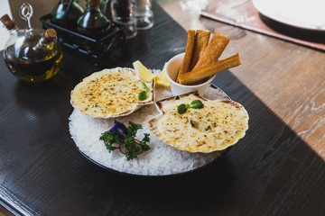 Baked scallops with cheese served with sliced lemon and bread sticks on bowl with full of sea salt.