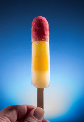 gefrorenes fruchtiges tropisches Eis am Stiel rot gelb weiß in der Hand blauer Hintergrund