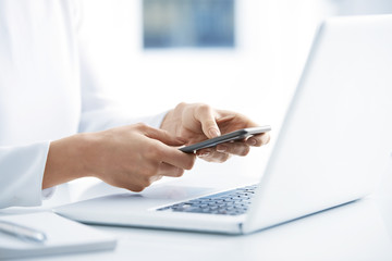 Fototapeta premium Woman using her mobile phone and send message. Close-up shot of businesswoman holding cellphone in her hands and text messaging while sitting at office desk in front of laptop. 
