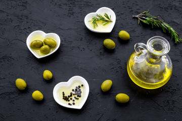 Fresh olive oil in glass jar near green olives and branch of rosemary on black background top view