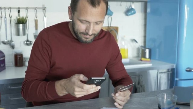 Young man paying bills on smartphone app by table at kitchen
