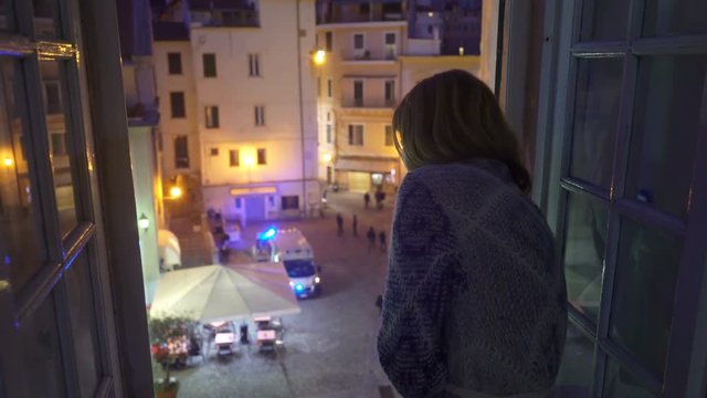 A night incident in the city. Ambulance car on the street. Excited Young woman looks out the window of her house. First aid, life insurance, terrorist threat. Home quarantine coronavirus Italy