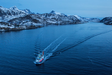 Fisher boat going for fishing before sunrise with mountains full of snow in background. Lofoten Island, Norway.