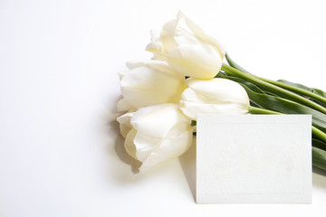 Feminine festive composition with bunch of white tulips, present wrapped in craft paper & blank greeting card. Beautiful bouquet of flowers for mother's day. Background, copy space, top view, close up