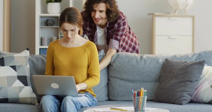 Young pretty woman in the yellow sweater and glasses sitting on the sofa and taping on the laptop computer when her boyfriend coming from behind, leaning on the couch and laughing. Indoors