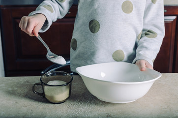 A baby in a grey sweater in the kitchen pours sugar out of the cup with a spoon into a large white porcelain bowl.