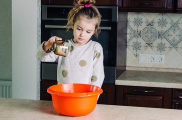 A pretty baby in a gray sweater in the kitchen pours sugar out of the cup into a large orange plastic bowl. © Olga