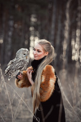 Woman blond in autumn in fur coat with owl on hand first snow. Beautiful girl with long hair in nature, holding an owl. Romantic, delicate look girls
