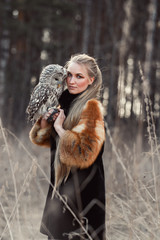 Woman blond in autumn in fur coat with owl on hand first snow. Beautiful girl with long hair in nature, holding an owl. Romantic, delicate look girls