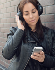 A beautiful girl in a black leather jacket listens to music from external headphones from her...