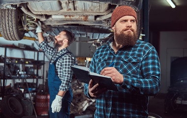 Two bearded brutal mechanics repair a car on a lift in the garage. 