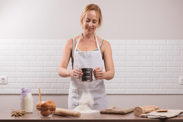 Young woman sifting flour through a sieve on  wooden table. The chef in white apron sifts the flour through a sieve to prepare the dough for pizza on a light background.