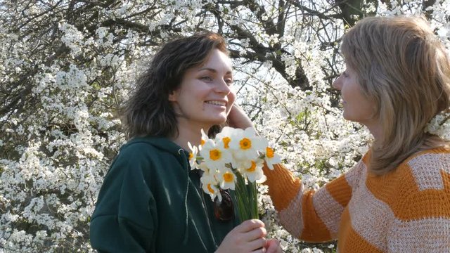Portrait of adult middle-aged mother and her adult daughter who gives a bouquet of daffodils for Mother's Day against the background of a flowering tree