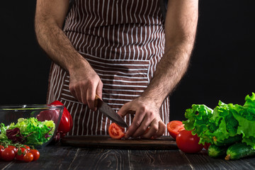 Man preparing salad on a wooden table. Men's hands cut the tomato to make a salad on black background