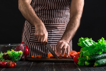Young home cook man in apron slicing carrot with kitchen knife.